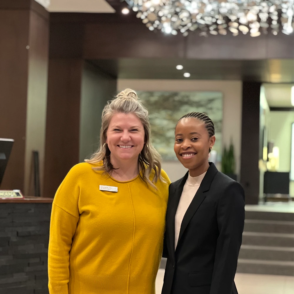 Two students stand, smiling and posing for a photo in a hotel lobby with a fancy chandelier.