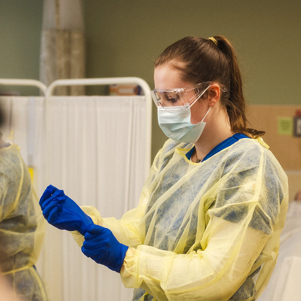 A nursing student puts on a medical smock and gloves over their scrubs.