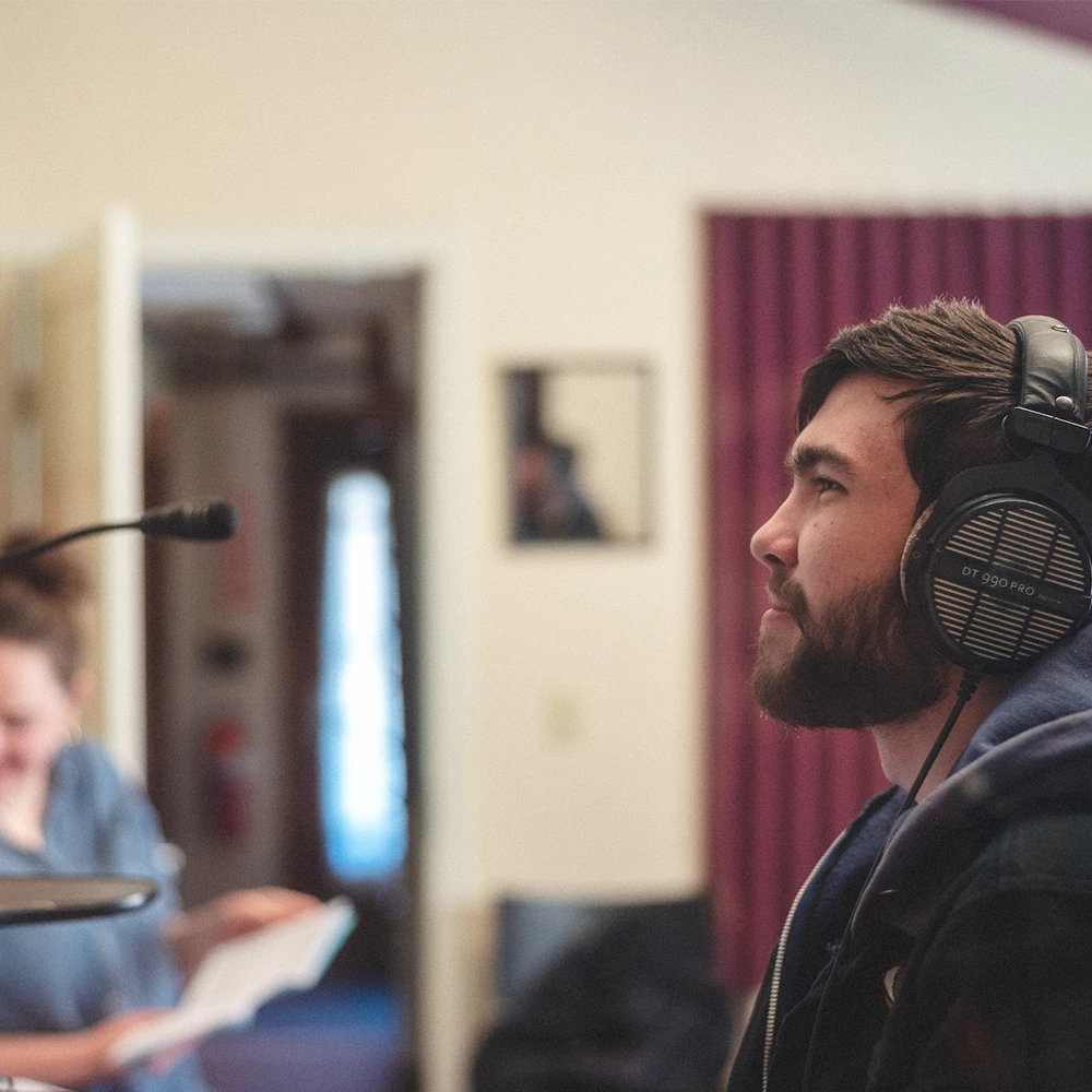 A student sits in a sound studio wearing headphones in front of a microphone.