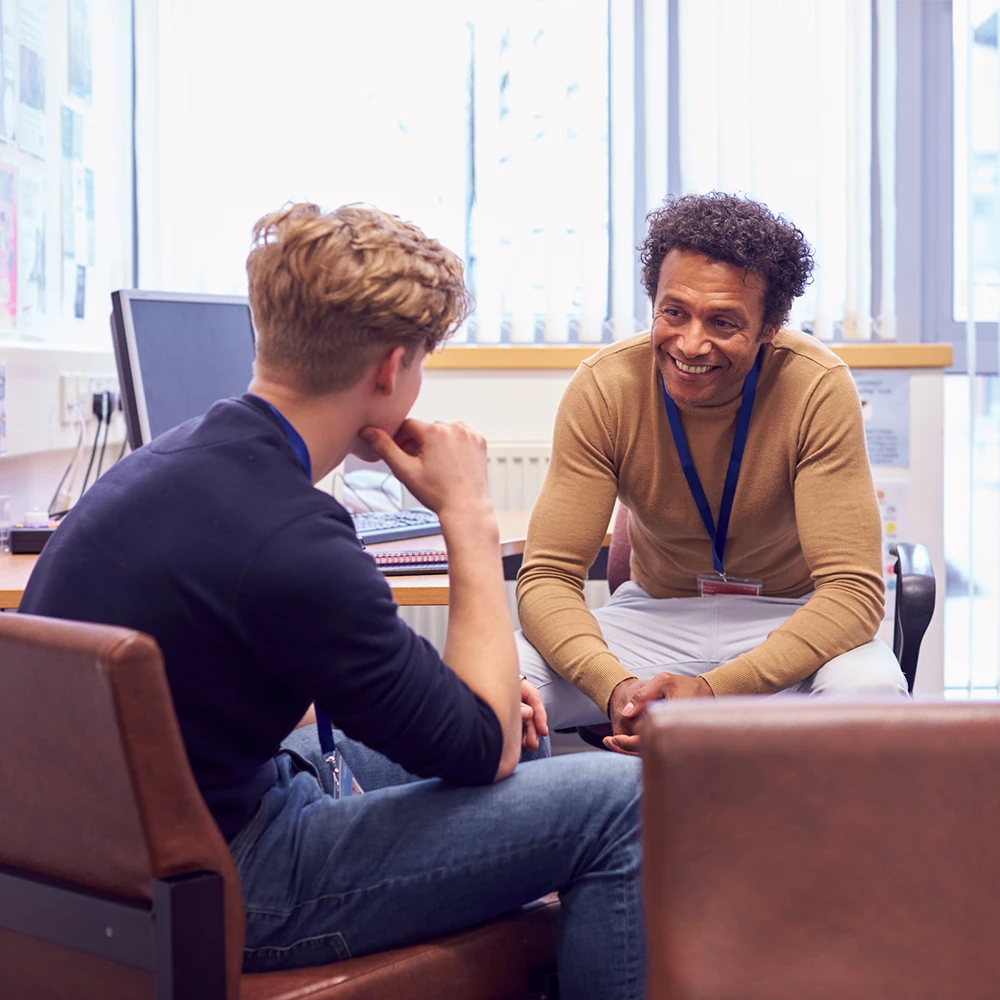 A school staff member wearing a badge sits in a chair and leans forward to talk with a student who is also sitting.