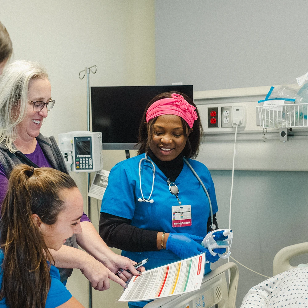 Nursing students look at a chart a faculty member is holding, standing next to a hospital bed a simulation lab.