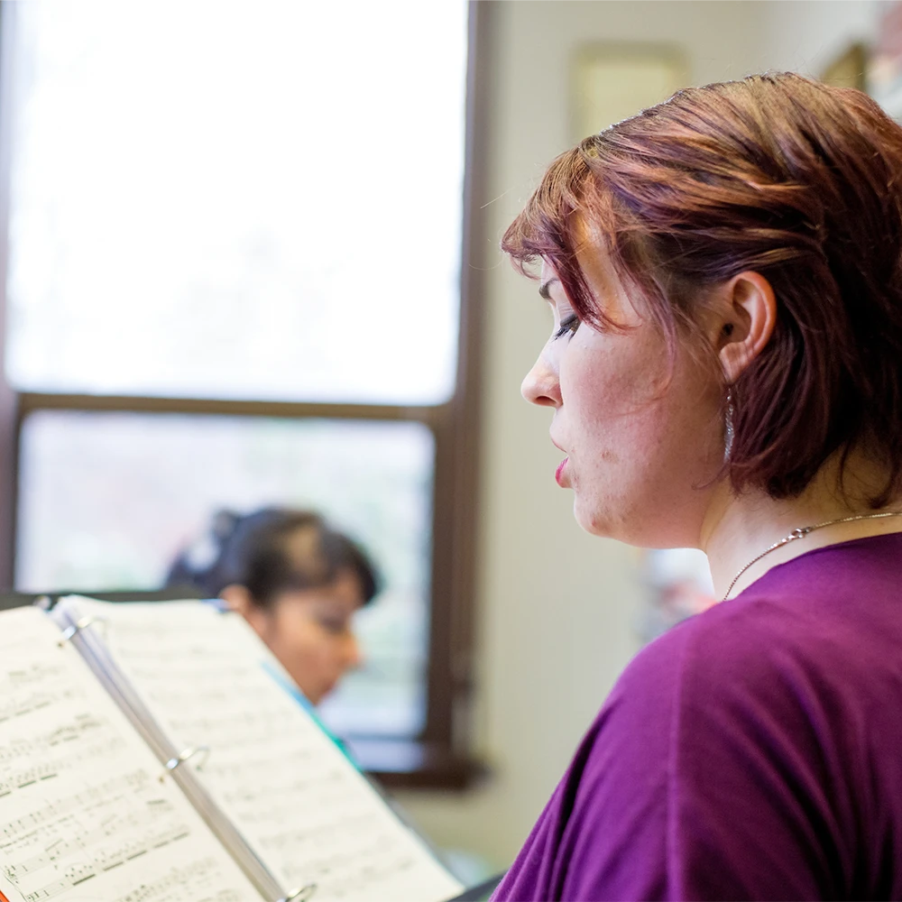 A student sings while reading music with a instructor playing piano in the background.