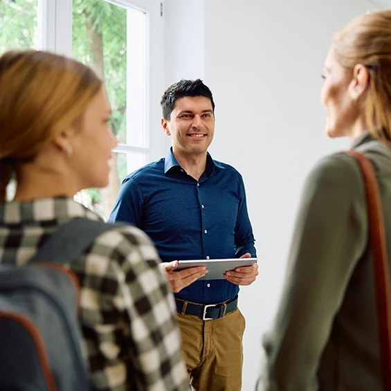 A school staff member stands in a hallway holding and ipad. He is looking at a mother and daughter. We see the back of their heads and the girl's backpack.