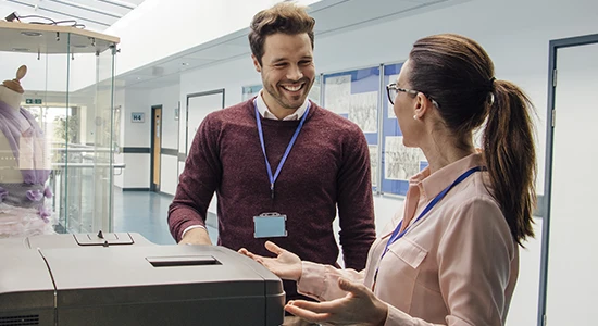 Two school staff members talk to each other in a hallway, standing by a photocopier.