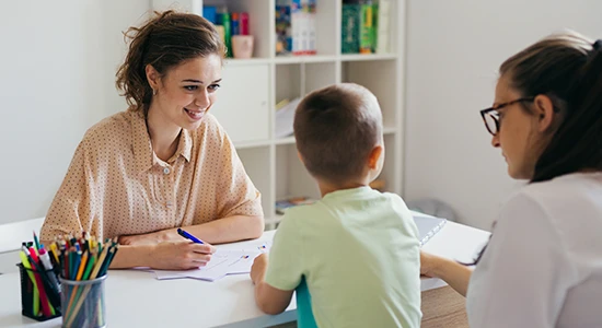 A school psychologist meets with a student and his parent. They sit at a table together with paper and a cup of pencils.