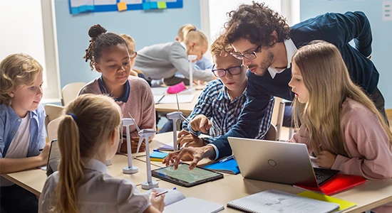 A teacher leans over a desk and works on an ipad, surrounding by elementary students and small model windmills.