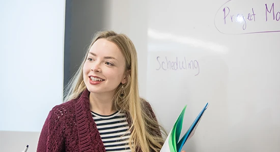 A student stands by a whiteboard and holds a folder.