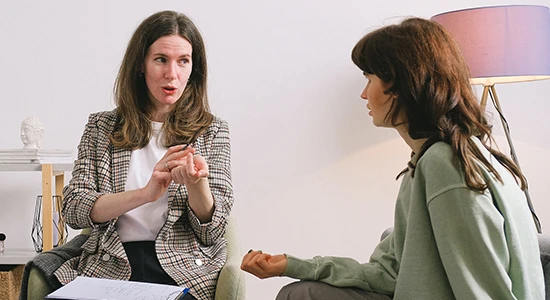 A counselor sits and gestures with her hands while talking to another seated woman who is listening.