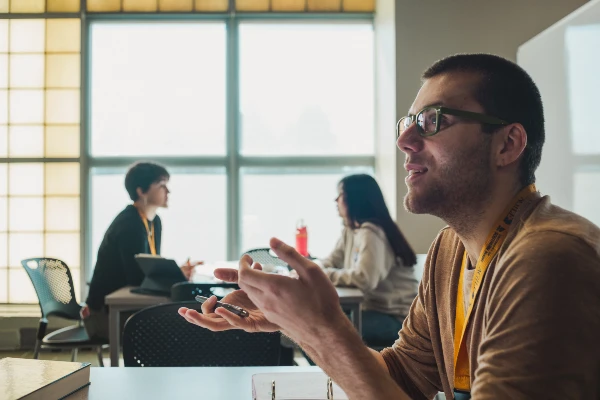 A student in the foreground engaged in conversation. Two students are seated at a table in the background.