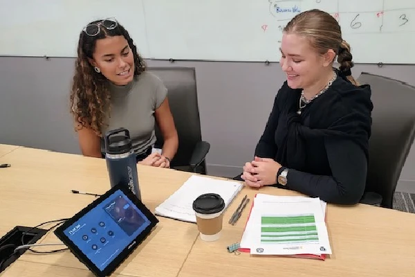An intern working at a table in front of a whiteboard with a mentor.