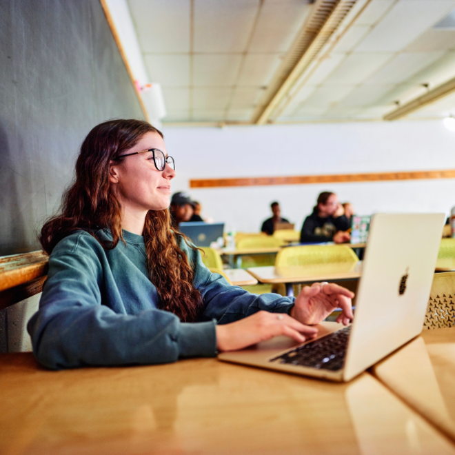 Student using a laptop in a classroom, seated in front of a chalkboard.