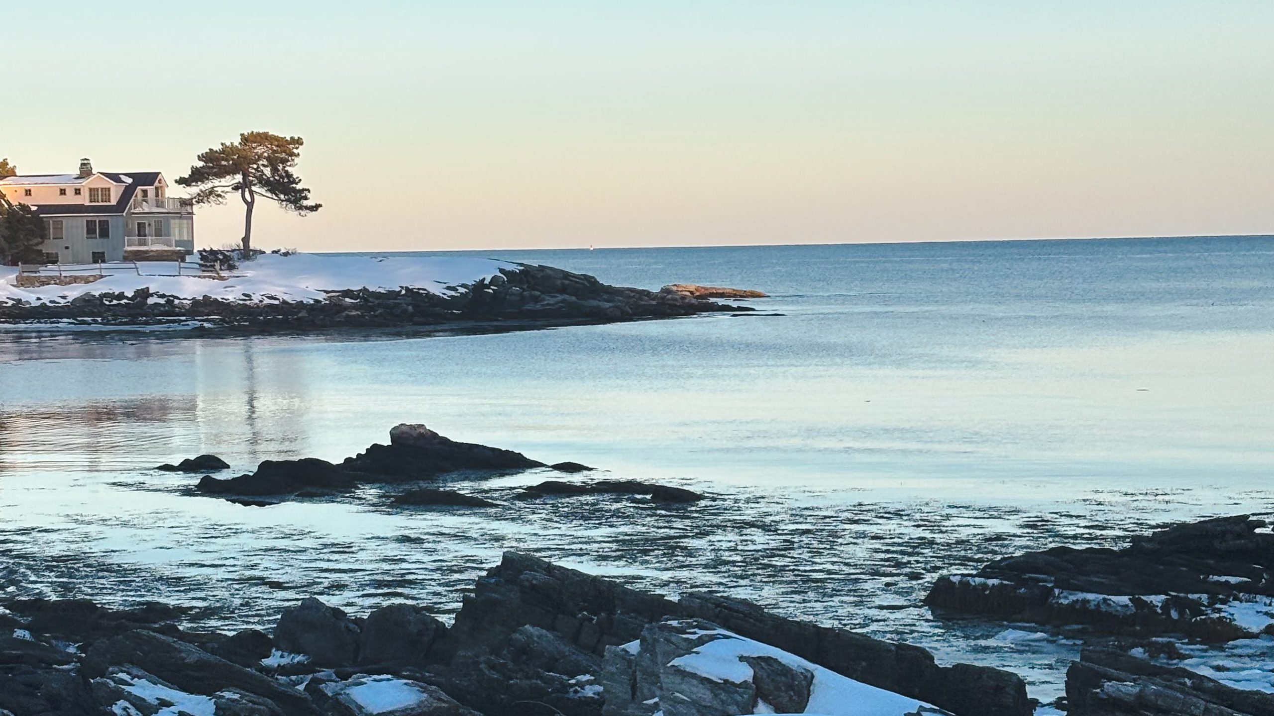Golden hour light over a snowy winter beach with glassy water and rocky shoreline in Southern Maine
