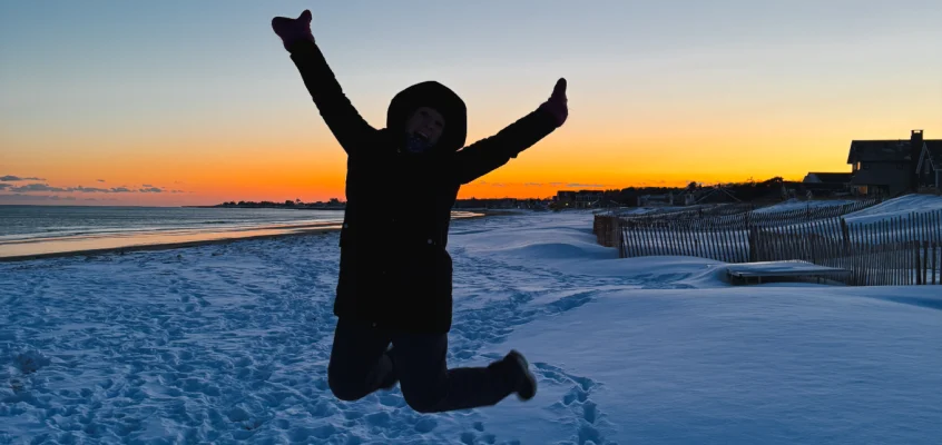 A person jumps with arms in the air on a snowy beach at sunset.