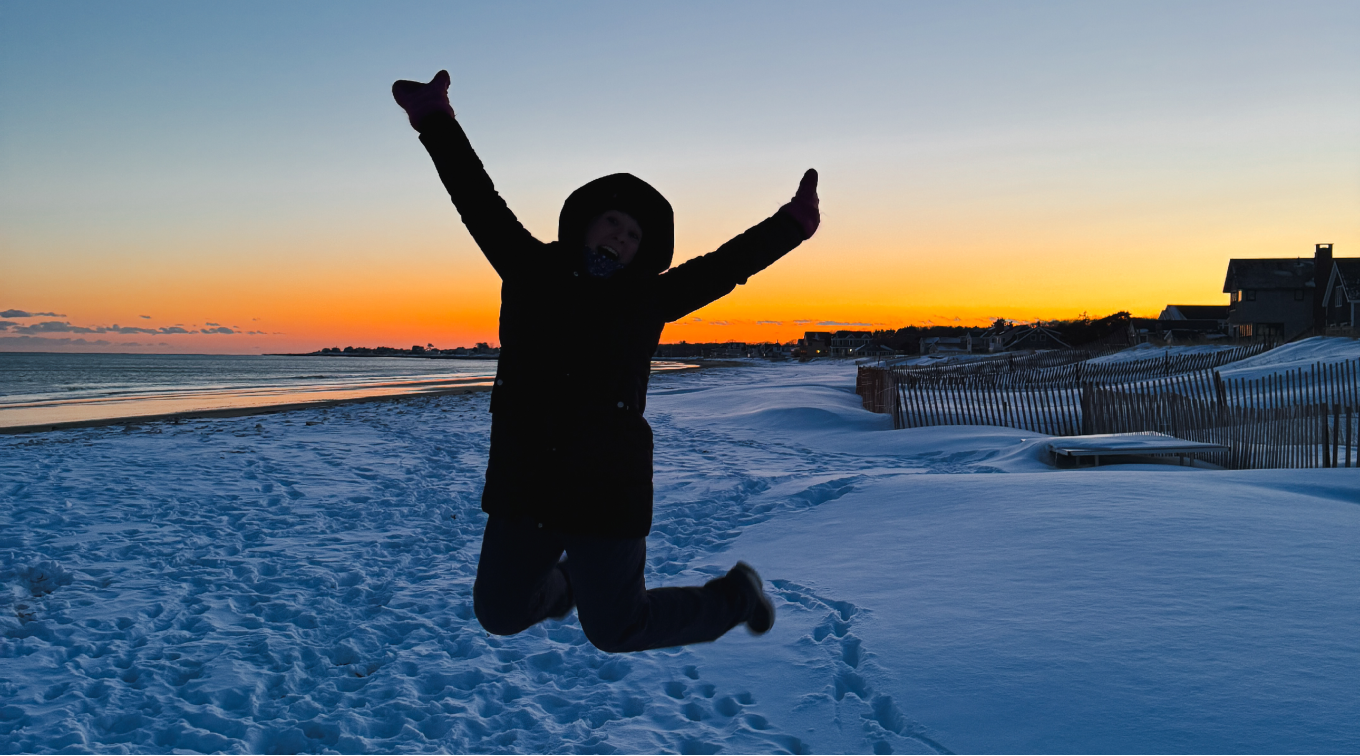 A person jumps with arms in the air on a snowy beach at sunset.