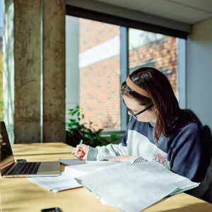 A student sits at a desk surrounded by windows, writing and studying.