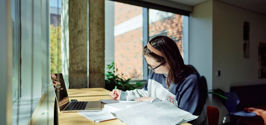 A student sits at a desk surrounded by windows, writing and studying.