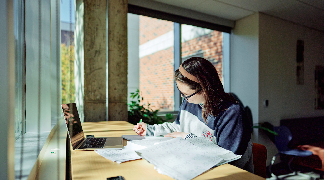A student sits at a desk surrounded by windows, writing and studying.