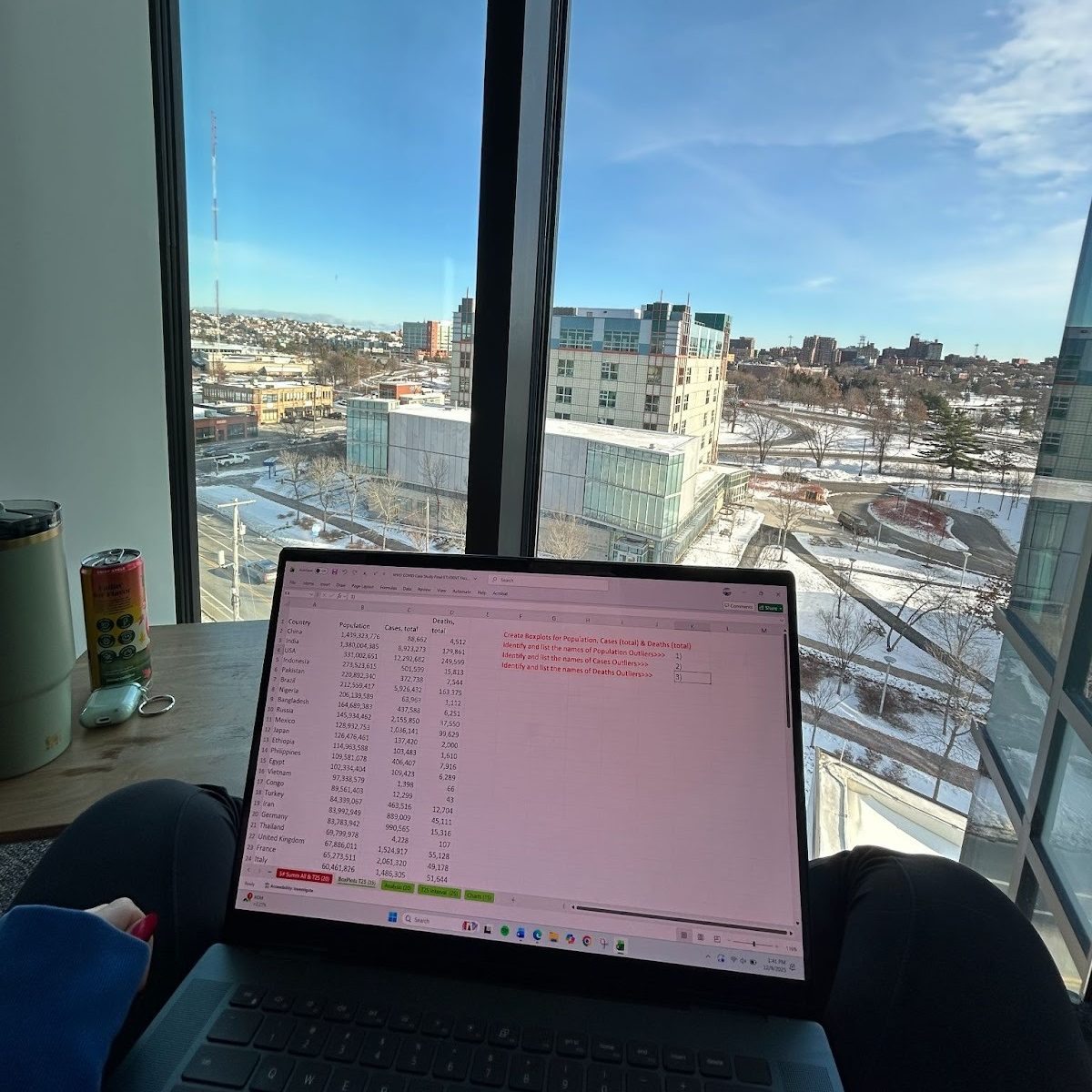 A student sits with a laptop resting on their lap, with large windows behind them overlooking the Portland, Maine skyline.