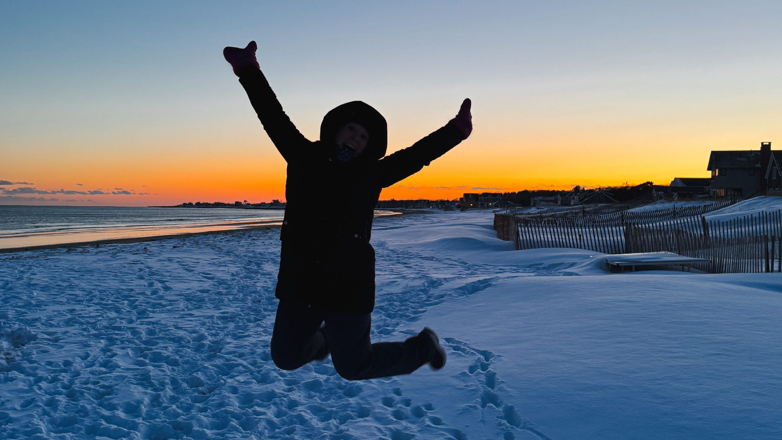 Person jumping with arms raised in celebration on a snowy beach at sunset in Southern Maine