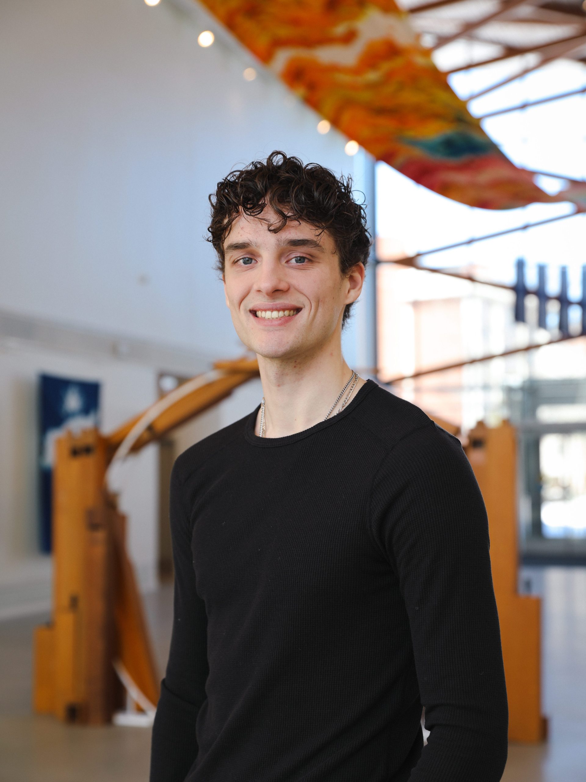 Vertical headshot of USM student Maddox Cahill looking at the camera in the Crewe Center Great Hall, with windows and artwork in the background.