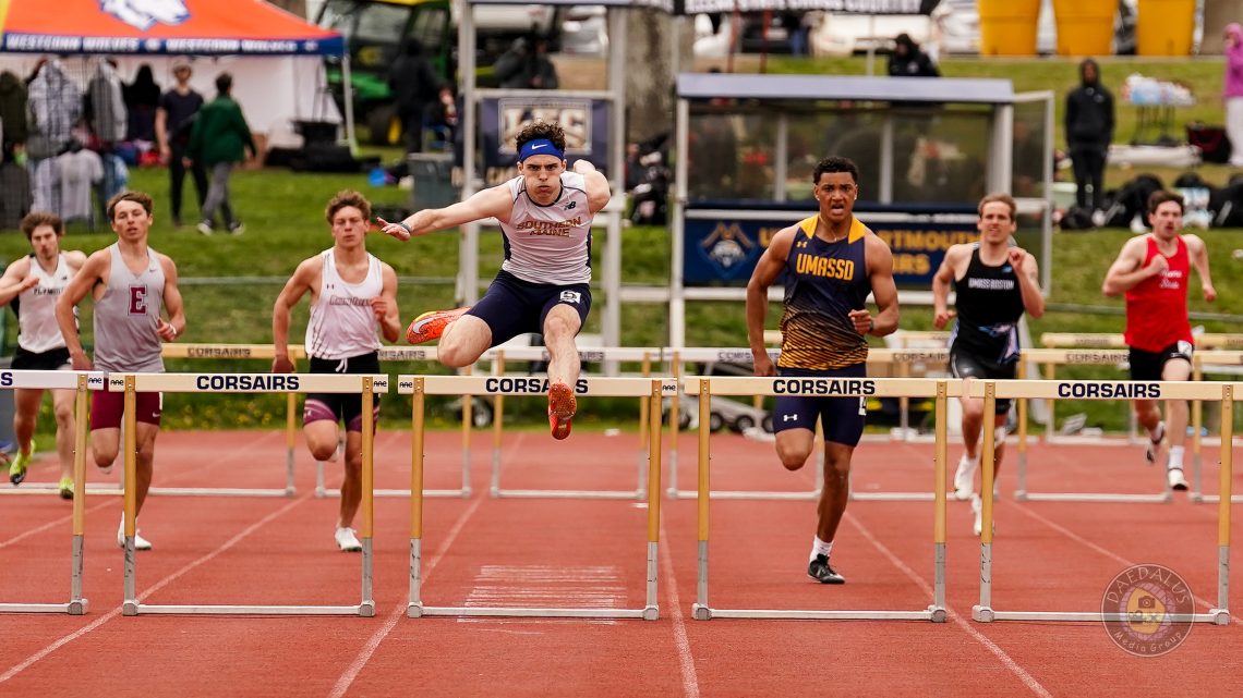 USM student Maddox Cahill clears a hurdle mid-jump during a track meet, centered in the frame with other runners on either side competing in adjacent lanes.