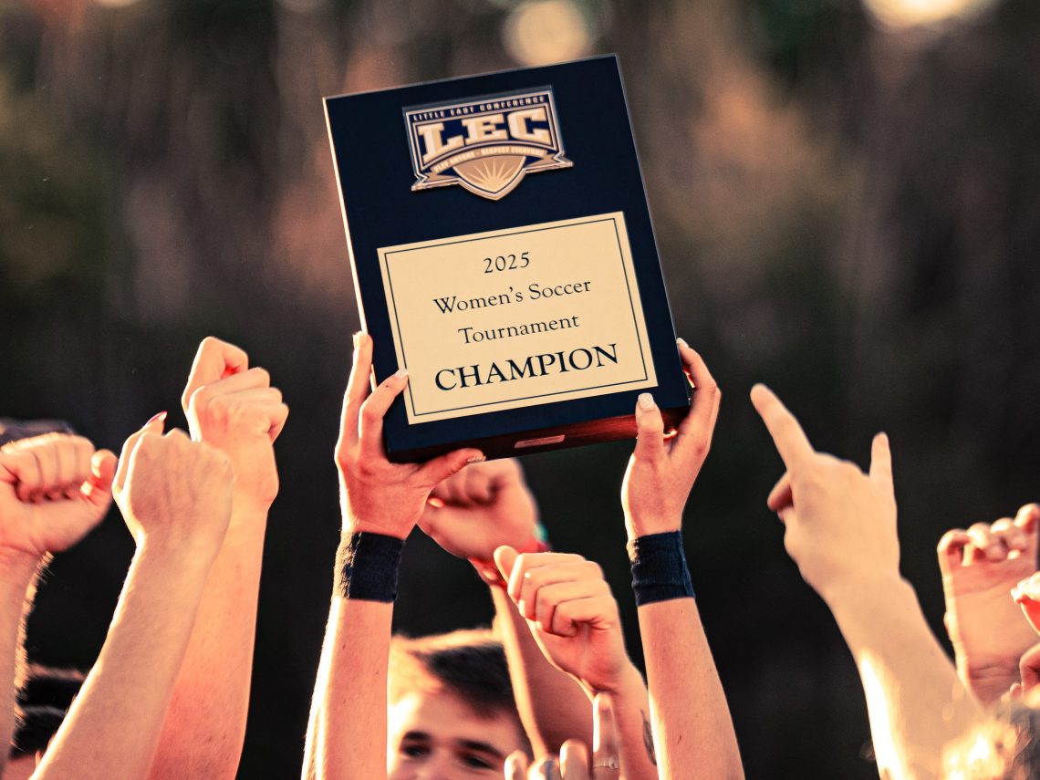 Close-up of USM women’s soccer players’ arms holding up their fourth Little East Conference championship trophy.