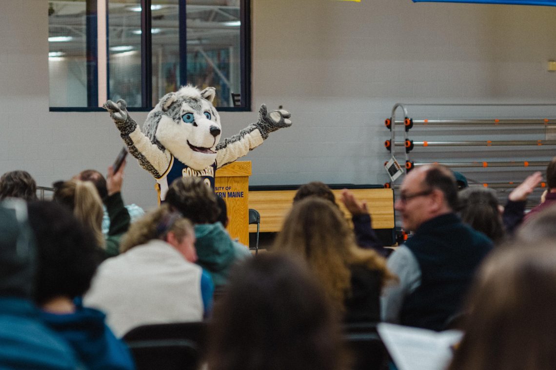 Champ the Husky mascot stands at a podium with arms raised in front of a crowd