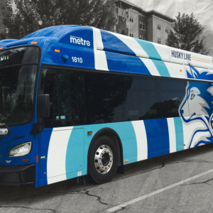 The University of Southern Maine's Husky Line bus stops on the street to pick up students. The background around the bus is black and white, and there are hand drawn yellow stars on either side of the bus.