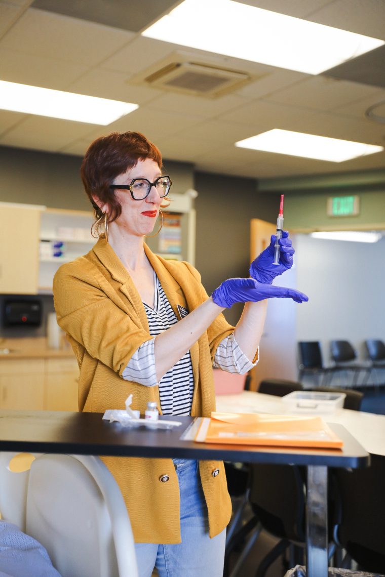 Christina Hart smiles while demonstrating how to draw medication into a syringe, holding the needle up in a nursing simulation lab.