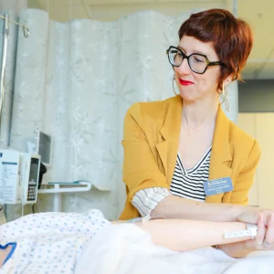 Christina Hart smiles while holding the hand of a simulation dummy, demonstrating patient care and compassion in the nursing lab.