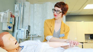 Christina Hart smiles while holding the hand of a simulation dummy, demonstrating patient care and compassion in the nursing lab.