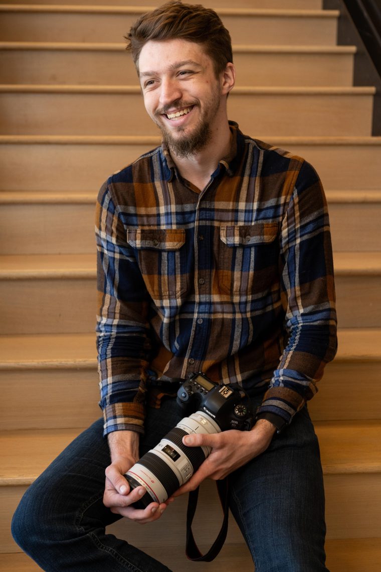 USM senior Deklin Fitzgerald sits on a staircase smiling, holding his camera.