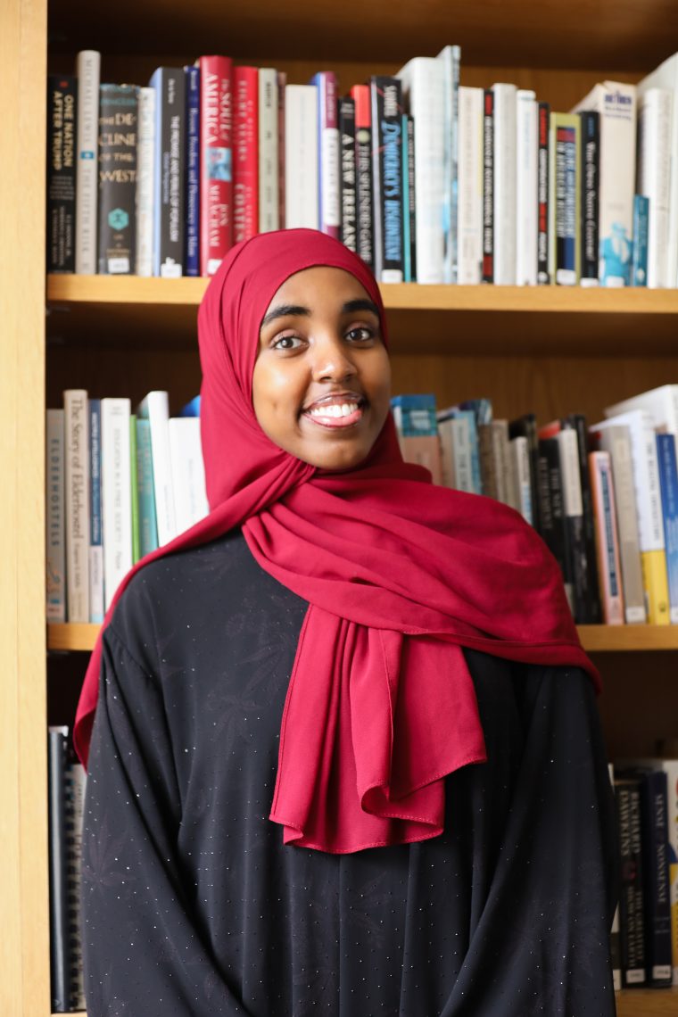 Najmo Ahmed smiles in front of a wooden bookshelf filled with books.