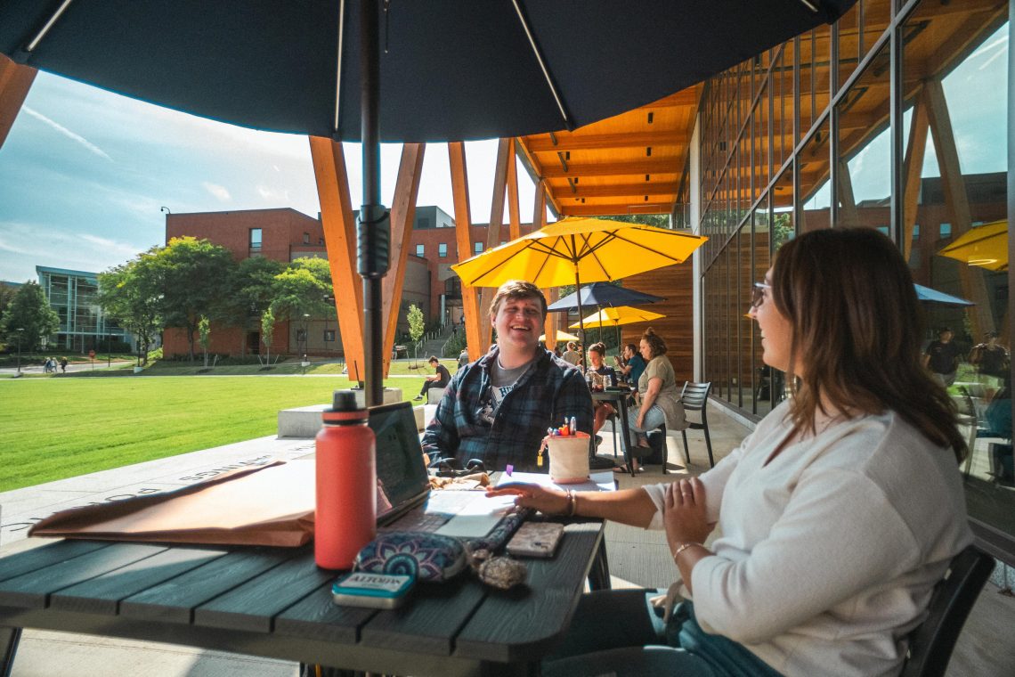 Students study at an outdoor table near McGoldrick Center on a sunny day, with green lawn and patio umbrellas in the background.