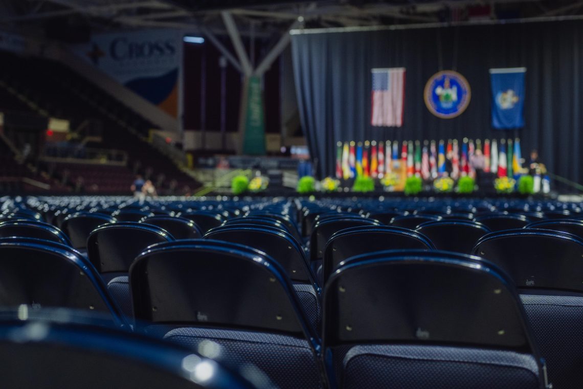 Empty seats face a decorated commencement stage adorned with flags and flowers.