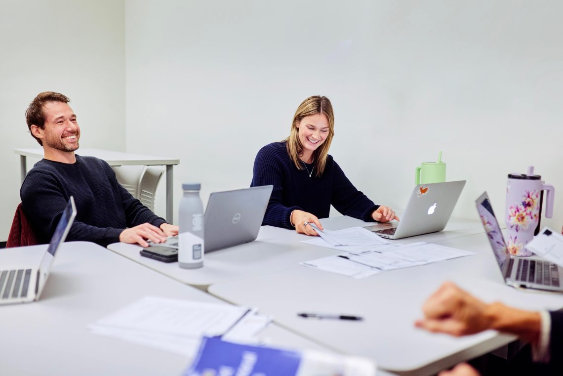 Two USM students smile and laugh during a group meeting, with laptops and papers spread across the conference table.