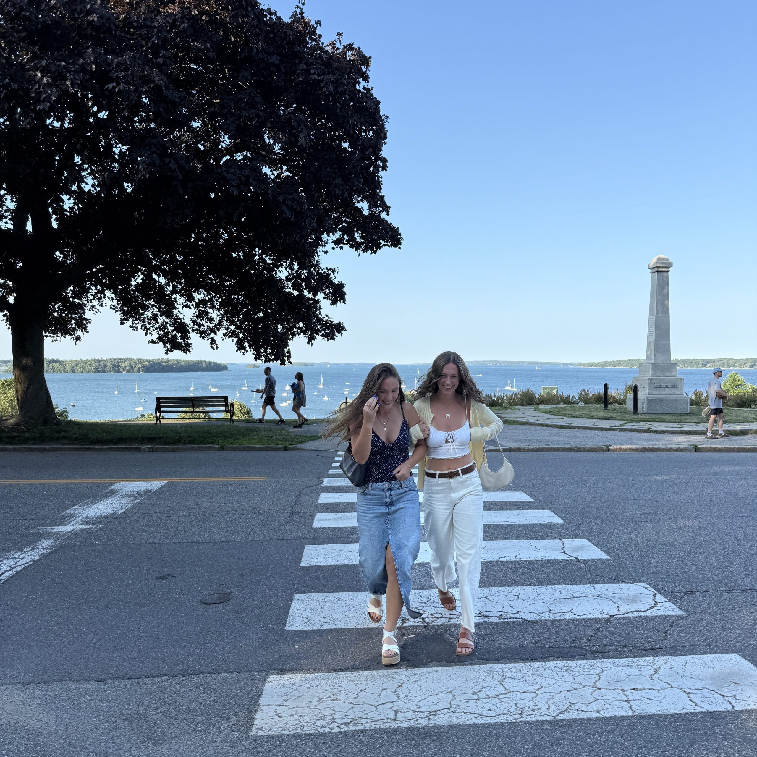 Bridgette and her friend walking across a street near the Eastern Promenade in Portland, Maine, with the ocean visible in the background.