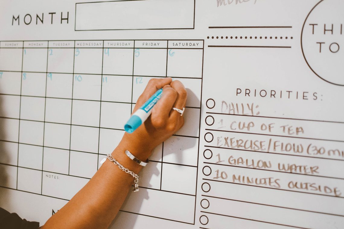 A person writes on a large wall calendar with a blue marker, with a priorities and daily checklist visible alongside the monthly grid.