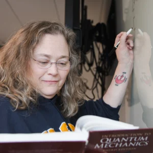 Lisa Struebing references a quantum mechanics textbook while writing an equation on a whiteboard in the Dubyak Center on USM's Portland campus.
