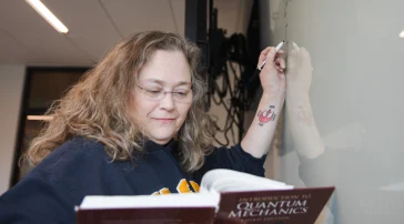 Lisa Struebing references a quantum mechanics textbook while writing an equation on a whiteboard in the Dubyak Center on USM's Portland campus.
