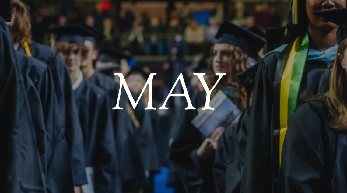 View down the aisle between rows of standing graduates in caps and gowns at USM commencement. The word "May" overlays the image.