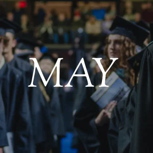 View down the aisle between rows of standing graduates in caps and gowns at USM commencement. The word "May" overlays the image.