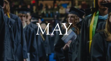 View down the aisle between rows of standing graduates in caps and gowns at USM commencement. The word "May" overlays the image.