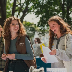 Two students smile and talk at an outdoor table on a sunny day during USM freshman orientation, holding papers in hand.