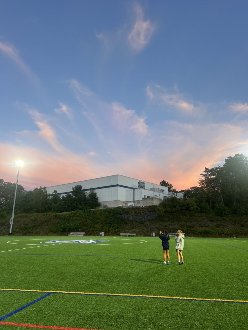 Two USM women's soccer players stand on the field in Gorham, photographing the sunset after practice.