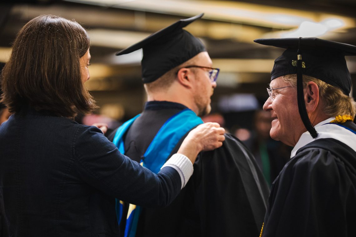 A faculty member adjusts the hood of a master's degree graduate's gown before the ceremony, with another graduate in cap and gown visible alongside them.