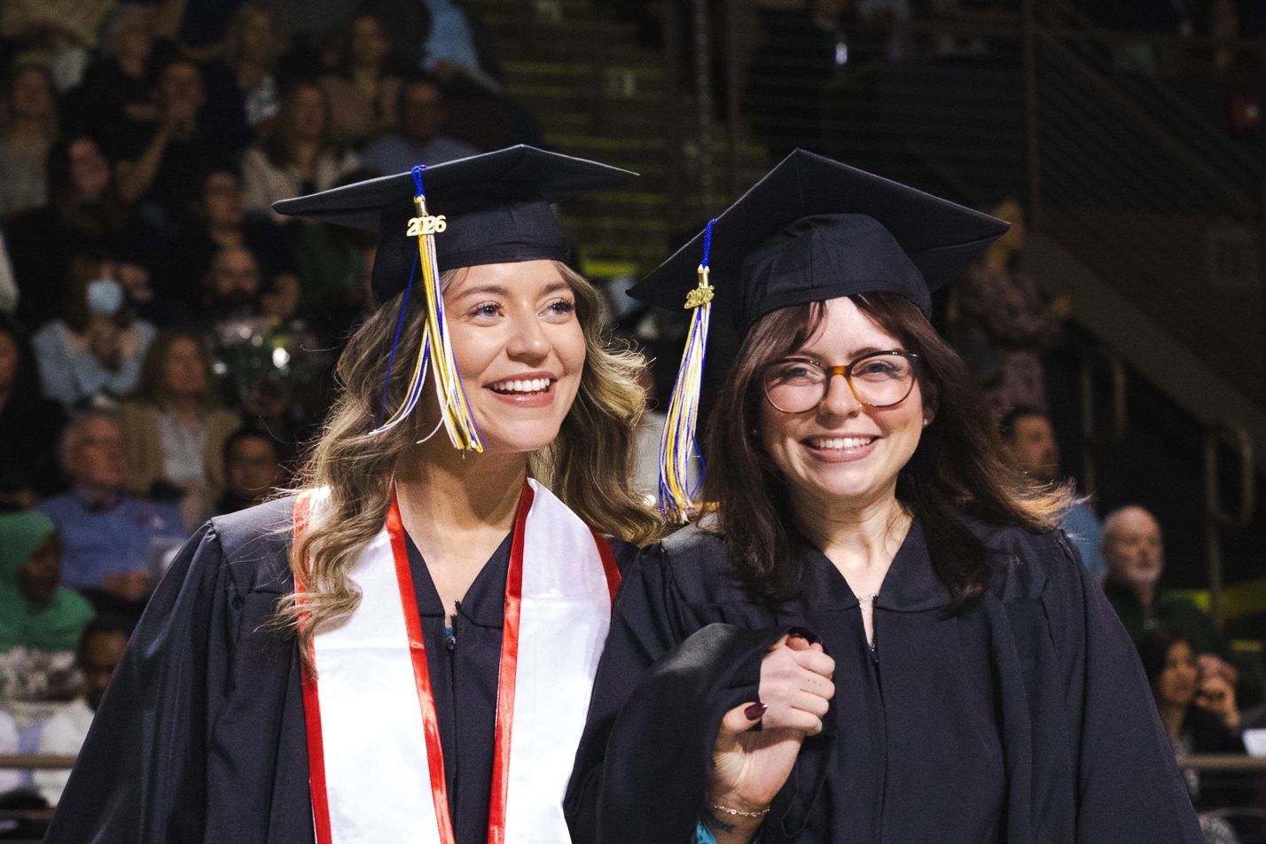 Two graduates in black caps and gowns smile at the camera as they cross the stage at Cross Insurance Arena, with a packed crowd visible in the stands behind them.