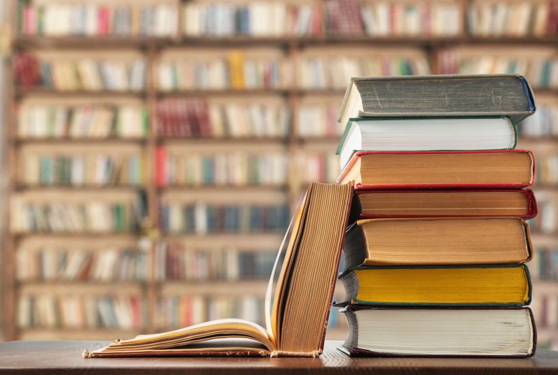 A stack of colorful books beside an open book on a wooden surface, with a blurred bookshelf in the background.