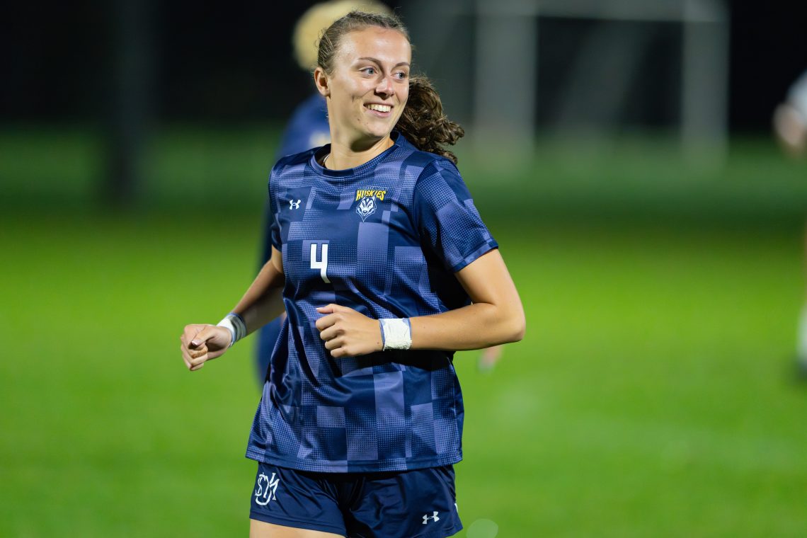 Bridgette Toland, wearing a USM Huskies navy jersey with the number 4, jogs on a soccer field during a nighttime game, smiling broadly.