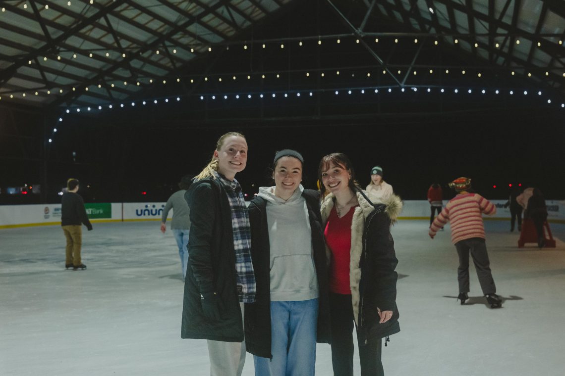 Three USM students smile together on an outdoor ice rink at night, with strings of warm lights overhead and other skaters in the background.